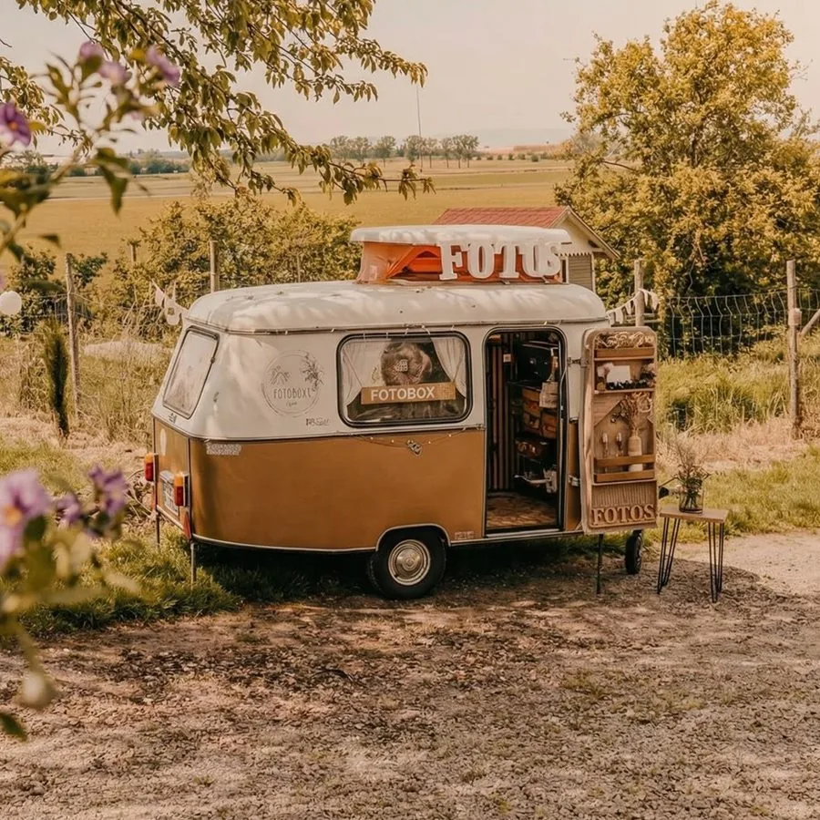 Der geöffnete Fotoanhänger am Feldrand, auf dem Dach der Leuchtschriftzug FOTOS, daneben ein Beistelltisch